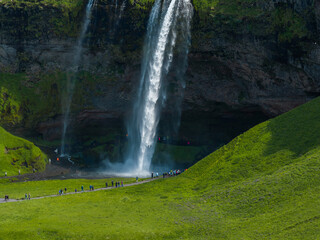 Seljalandsfoss waterfall cascades down a rocky cliff into a pool while tourists walk along the footpath below in the grassy landscape of southern Iceland. © Aerial Film Studio