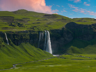 Seljalandsfoss waterfall cascades from grassy cliffs onto the plains below in Iceland. Tourists walk along a pathway winding through the meadows near the base of the cascade. © Aerial Film Studio