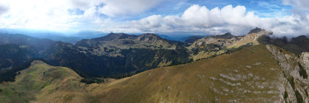 Aerial view of the Pointe d'Uble mountain range with rugged slopes, green alpine meadows, dense coniferous forests, and scattered clouds in the sky in Taninges, Auvergne-Rhone-Alpes, France.