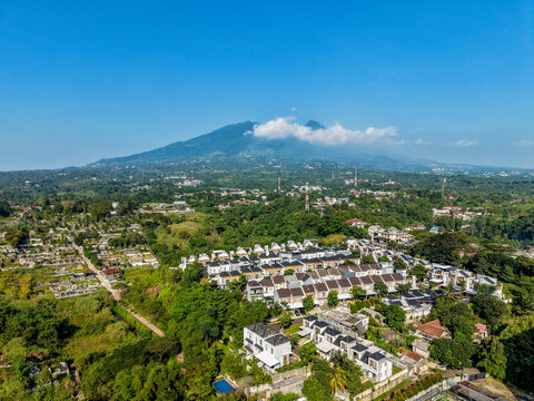 Aerial view of a residential neighborhood with modern houses nestled among lush green trees, with the majestic Mount Salak rising in the background in Bogor City, West Java, Indonesia.