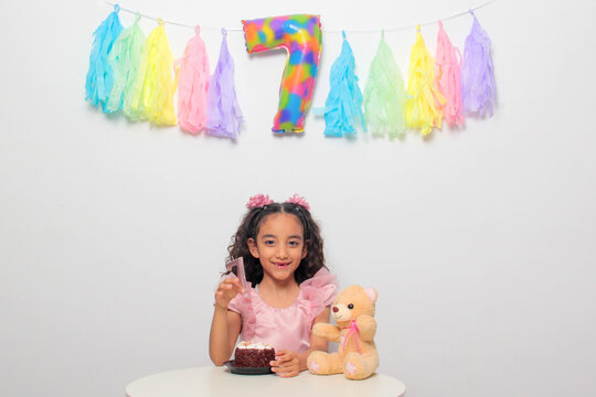 Seven-year-old Latina girl with brown skin and curly hair celebrates her birthday with cake, candle, and colorful decorations