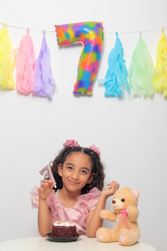Seven-year-old Latina girl with brown skin and curly hair celebrates her birthday with cake, candle, and colorful decorations