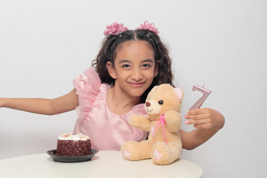 Seven-year-old Latina girl with brown skin and curly hair celebrates her birthday with cake, candle, and colorful decorations