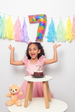 Seven-year-old Latina girl with brown skin and curly hair celebrates her birthday with cake, candle, and colorful decorations