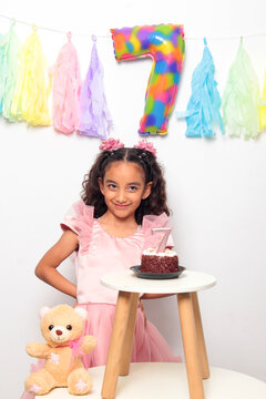 Seven-year-old Latina girl with brown skin and curly hair celebrates her birthday with cake, candle, and colorful decorations