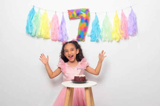 Seven-year-old Latina girl with brown skin and curly hair celebrates her birthday with cake, candle, and colorful decorations
