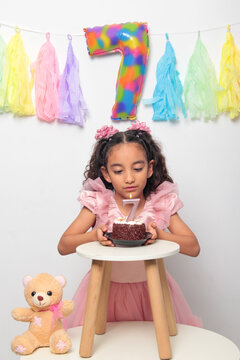 Seven-year-old Latina girl with brown skin and curly hair celebrates her birthday with cake, candle, and colorful decorations