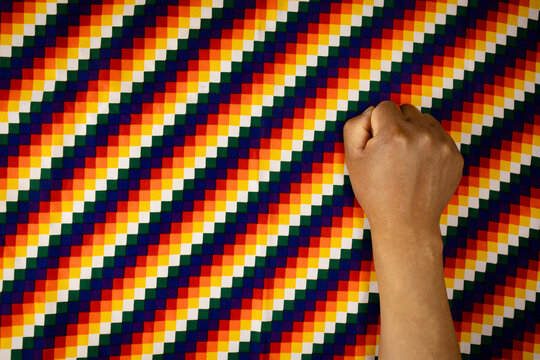Hand of an indigenous man from the Andean region of South America, against a Wiphala background.