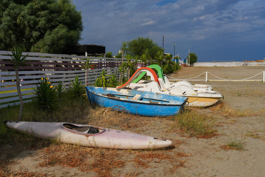 Old pedal boats with slides, blue rowing boat and kayak stored on empty sandy beach after end of summer season, Calabria, Italy