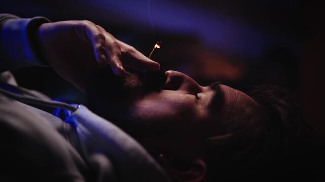 A Caucasian man reclines in dim blue light and smokes a marijuana joint, bringing it to his mouth and exhaling. Cannabis use and addiction concept.