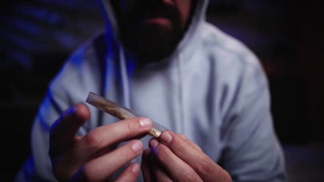 A Caucasian man in a light hoodie prepares a marijuana joint with rolling paper in a dark room. Cannabis use and addiction concept.