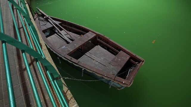 Old weathered wooden rowing boat moored with a thin chain on dark green lake water beside a teal metal railing and paved walkway at night, overhead view, two oars inside, urban park waterfront concept