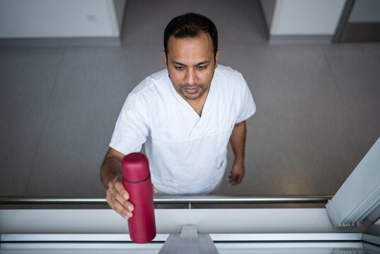 High angle view of a male healthcare worker in white scrubs reaching for a red thermos bottle on a reception counter in a hospital