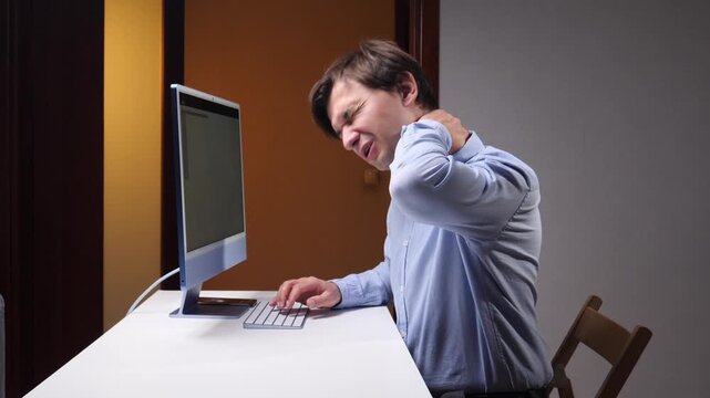 Office worker gripping his neck while using a desktop computer, experiencing muscle pain and work related strain at a desk in a quiet indoor workspace