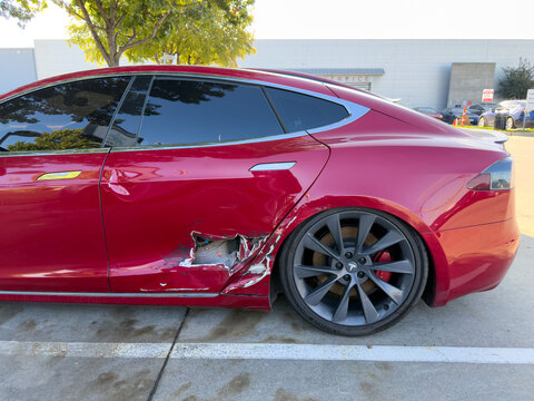 Dallas, Texas, USA - October 15th, 2026: Damaged red Tesla Model S sedan parked in service center lot with severe side door impact collision wear