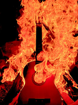 An acoustic guitar burning in a fire at night, with the guitar's body and neck visible amid intense orange flames.