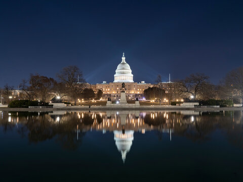 The illuminated U.S. Capitol dome and facade reflected on wet pavement at night, with bare trees framing the scene under a clear night sky.