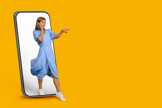 Smiling Young Woman Pointing At Smartphone Screen Inside Smartphone Frame With Yellow Background, Displaying Excitement And Engaging In Interaction, Mockup Concept.