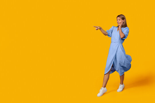 Smiling Young Woman In Blue Dress Pointing With Excitement At Something Off-camera In Studio With Bright Yellow Background, Full Of Energy And Positivity. Lifestyle Concept.