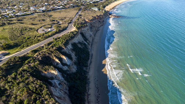 Aerial drone view of dramatic ocean cliffs along the coast of the rural town Anglesea on the Great Ocean Road in Victoria, Australia, with crashing waves on a sandy beach below, native coastal scrub.