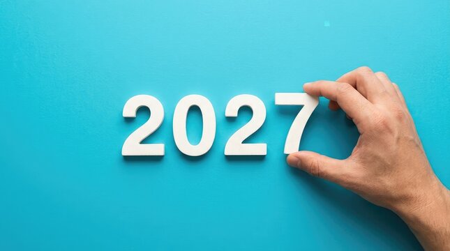 Hand adjusting wooden block year display on blue background with solid blue backdrop 2027 against solid block letters moves wooden future year