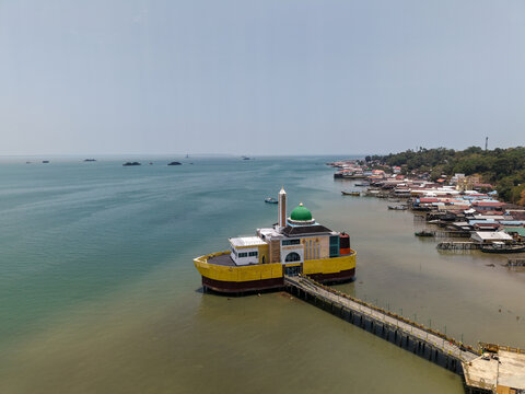 Aerial view of Masjid Apung Siring Laut Kotabaru, a floating mosque with a green dome and yellow hull, connected by a long pier to the coast of Kotabaru Regency, South Kalimantan, Indonesia.