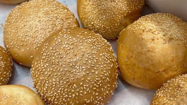 Fresh sesame burger buns on baking paper inside bakery shop display. Golden baked bread rolls offer artisan baking theme, fast food ingredient showcase, culinary preparation product.