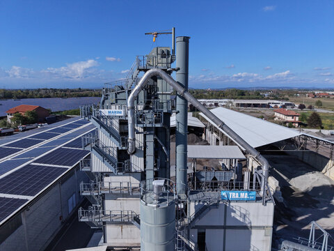 Aerial view of an industrial asphalt plant with tall metal silos, conveyor pipes, and solar panels on the adjacent warehouse roof in Torrazza Piemonte, Piemonte, Italy.