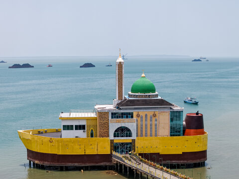 Aerial view of Masjid Apung Siring Laut Kotabaru, a unique mosque built on a yellow ship-shaped structure in the sea, connected by a pier in Kotabaru Regency, South Kalimantan, Indonesia.