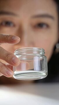 Vertical video: Hand entering, woman grasping lifting glass jar on table showing liquid to camera