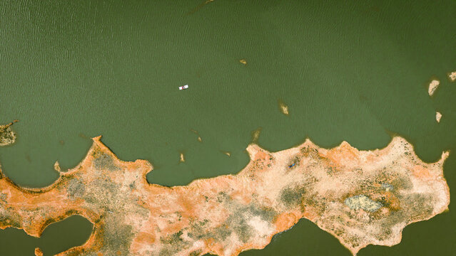 Aerial view of a small boat floating on the calm green waters of a lake with an irregular sandy shoreline in India.