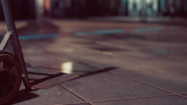 A close view of a floor with a shiny finish, capturing reflections of nearby objects. A cart with wheels rests at the edge, hinting at activity in a bustling area, perhaps a market or arena.