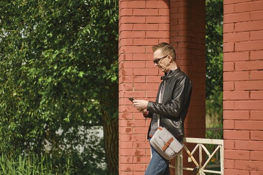 A man wearing sunglasses and a leather jacket stands by red brick columns, attentively using his smartphone. Lush green foliage surrounds the structure, indicating a pleasant sunny day