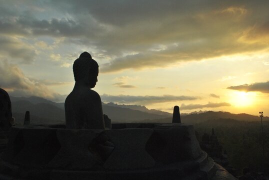Silhouette of an ancient stone Buddha statue at Borobudur temple, Indonesia during a beautiful golden sunrise. Majestic mountain landscape background. Peaceful spiritual travel destination.   