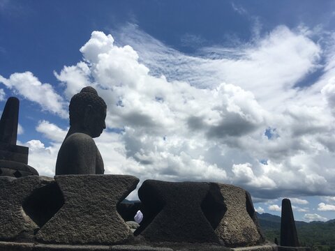 Side profile of an ancient stone Buddha statue at Borobudur temple, Indonesia. Majestic view against a bright blue sky with beautiful dramatic white clouds. Serene spiritual travel destination.   