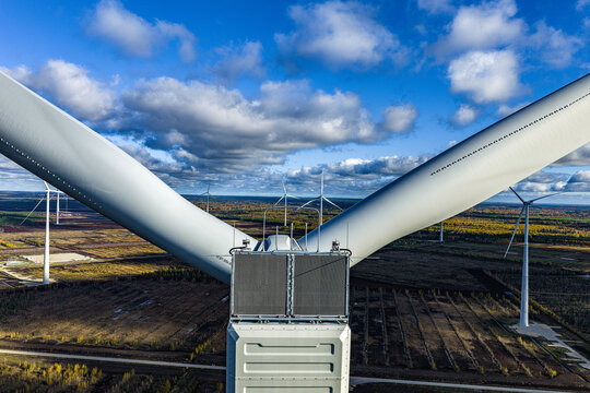 Aerial view of a wind turbine with large blades extending outwards, set against a backdrop of a vast wind farm, autumn forest, and blue sky in Jelgava, Latvia.