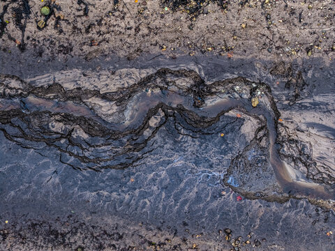 Aerial view of a winding muddy stream bed with intricate soil textures, dark wet earth, and natural sediment patterns in Jelgava, Latvia.