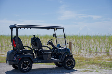An electric golf cart parked beside sand dunes in Florida and sea grass on a sunny beach, evoking summer leisure, coastal travel and relaxed vacation vibes for outdoor recreation and resort transport. © Del Harper