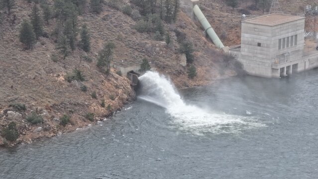 Aerial view of water discharging from a concrete dam outlet into a reservoir surrounded by rocky hillsides and pine trees in Estes Park, Colorado, United States.
