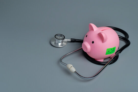 Piggy bank with an HSA green label encircled by a medical stethoscope with copy space. Health savings accounts, healthcare costs, medical insurance concept.