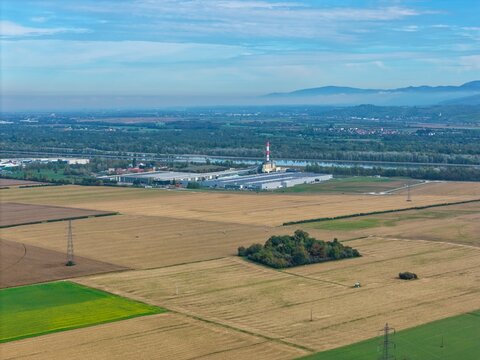 Aerial view of an industrial facility with a tall chimney, surrounded by agricultural fields, a river, and distant mountains in Hombourg, Grand Est, France.