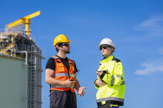 Male and female industrial engineers talking during site inspection near gas storage tank plant, Technician man holding walkie talkie discussing project with female colleague at refinery plant