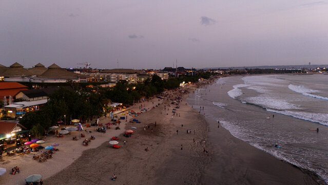 Aerial view of Kuta Beach at dusk, featuring the sandy shoreline, gentle waves, beach umbrellas, and nearby resort buildings in Kuta, Bali, Indonesia.
