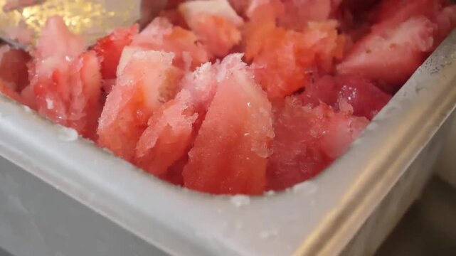 High angle, close up shot of a metallic spoon digging into and scooping a vibrantly red, freshly crushed strawberry shaved ice dessert (or watermelon granita) stored in a commercial stainless steel ki