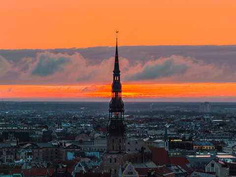 Aerial view of St. Peter's Church spire rising above the historic rooftops of Riga at sunset with a vibrant orange sky in Riga, Latvia.