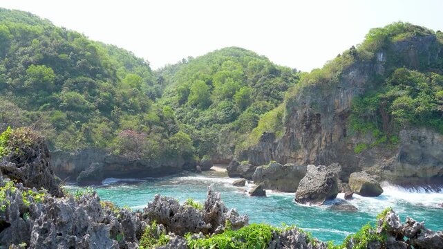 Scenic tropical coastal view with rugged rock cliffs covered in vegetation meeting vibrant blue ocean waves under a clear sky in Indonesia.
