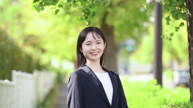 春の新緑が美しい屋外を笑顔で微笑む、新卒の若い日本人女性社員、大学生の就活や新入社員としてフレッシュなイメージのカメラ目線笑顔の動画