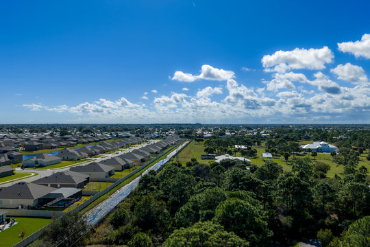 Aerial view of a suburban neighborhood featuring rows of houses, a drainage canal, and lush green trees under a bright blue sky in Grant-Valkaria, Florida, United States.