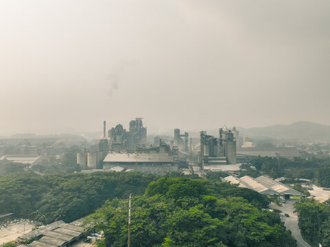 Aerial view of a large industrial cement factory complex with tall smokestacks and storage silos surrounded by dense green forest in Klapanunggal, West Java, Indonesia.