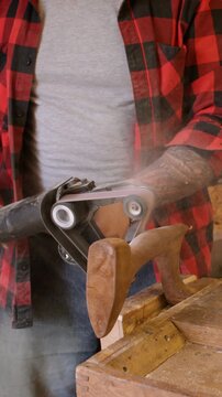 Vertical video. Carpenter equipment. Sanding wood. Man polishing figure piece product with handheld belt sander in joinery workshop spreading sawdust.